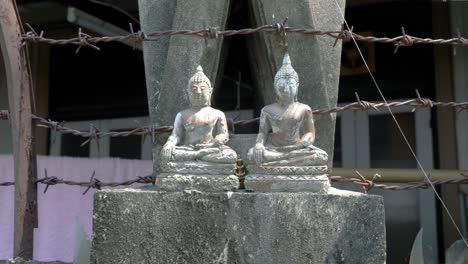 Two-small-stone-statues-of-sitting-Buddha-in-meditation-with-barbed-wire-in-background