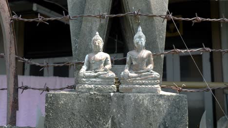 two small stone statues of sitting buddha in meditation with barbed wire in background