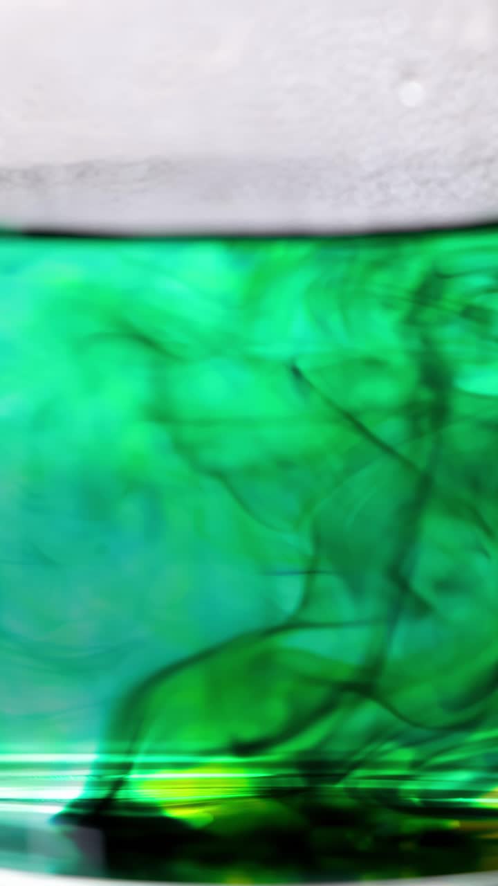 Colorante alimentario verde se arremolina y se dispersa en agua caliente, capturado en un recipiente de vidrio transparente.