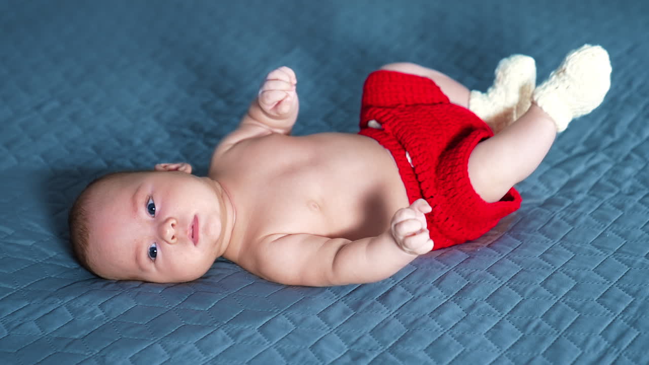 Blue-eyed Caucasian baby boy wearing red shorts and socks only tosses little feet. Beautiful child lying on the grey background.