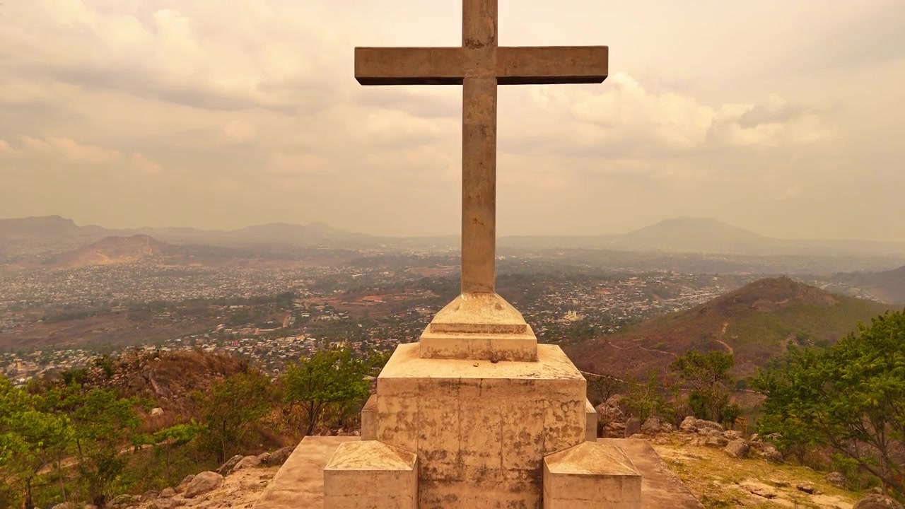 Drone rises from the base of the summit cross on Michiru Mountain, gradually revealing the full cross overlooking Blantyre from the Way of the Cross pilgrimage hike beneath a hazy sky.