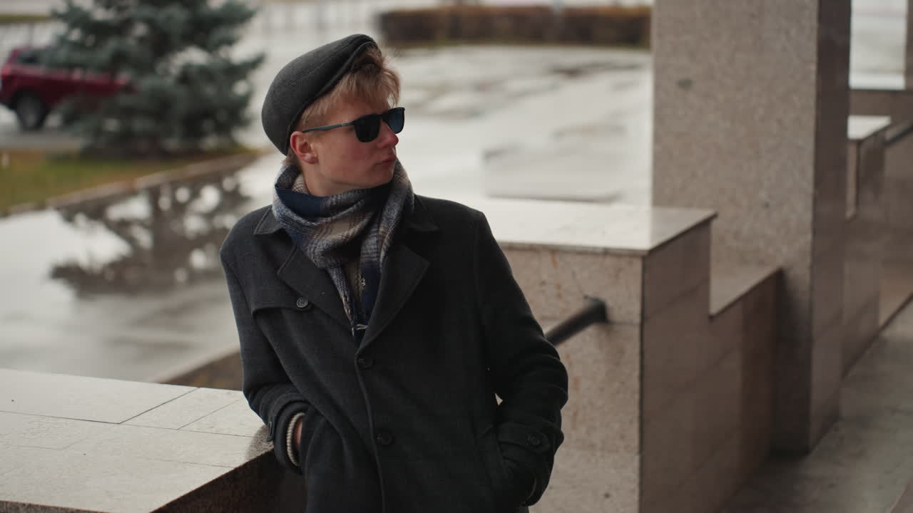 Elegante creatore bianco appoggiato a una ringhiera che si affaccia su una piazza cittadina piovosa, mani in tasca con occhiali da sole e cappotto a strati, pozzanghere riflettenti e toni smorzati, scena di moda urbana cinematografica perfetta