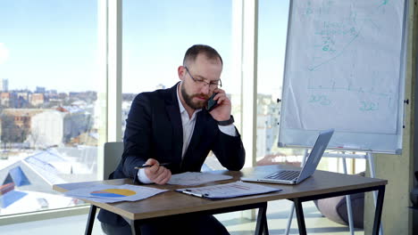 business worker at his workplace. confident man in glasses sitting at the desk with a laptop and talking on a phone looking into papers in office.