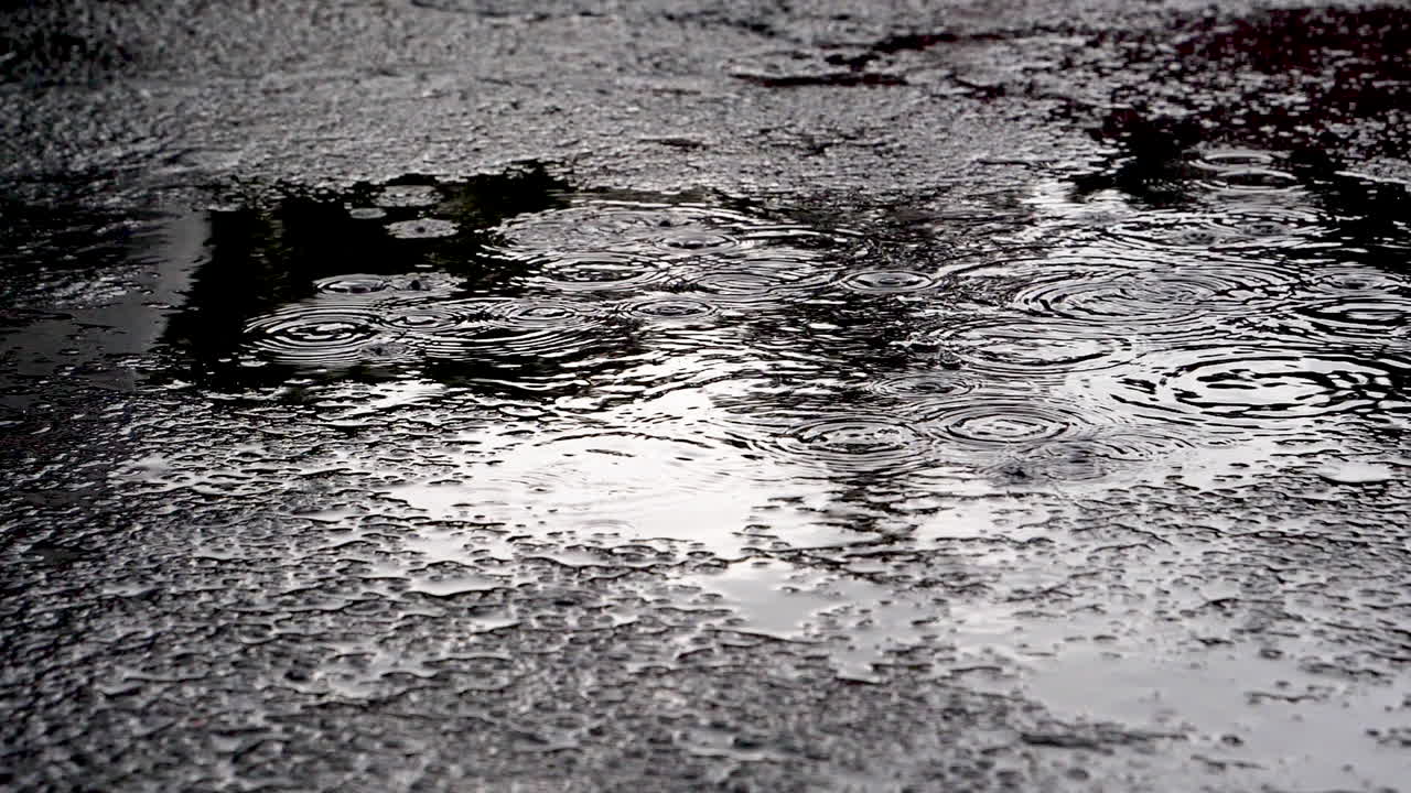 On a dark afternoon a sudden rain shower disturbs the quiet puddle located in a lonely parking lot