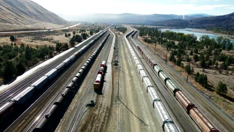 backwards-flight-dolly-drone-shot-flying-over-a-railroad-station-in-a-desert-environment-on-a-sunny-day-next-to-a-highway-and-a-river-and-mountains-in-the-background-and-powerines-in-the-picture