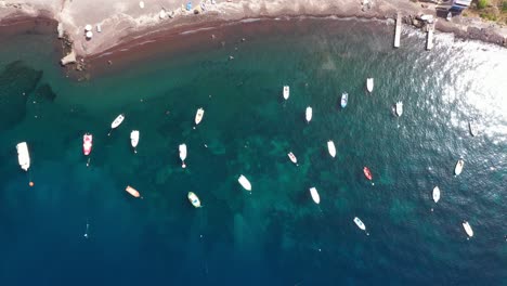 Vista-Aérea-De-Arriba-Hacia-Abajo-Que-Desciende-Sobre-Muchos-Barcos-De-Pesca-En-El-Mar-Mediterráneo-Con-Agua-Azul-Cristalina-Y-Playa-En-Santorini,-Grecia