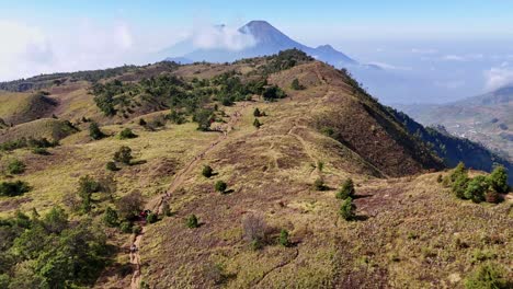 Drone footage of mountain hiking trail winding through grassy hills and trees, with stunning mountain peak rising in the distance. Mount Prau, Indonesia