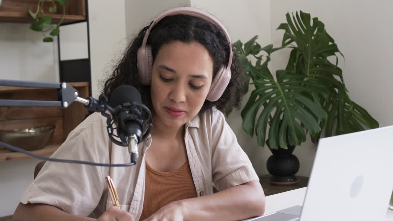 Mujer afroamericana con auriculares ajustando la mesa de mezclas, hablando por el micrófono y escribiendo notas para su podcast en el escritorio.