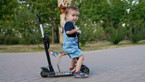 smiling kid in a jeans romper sitting on the scooter. happy baby pushes from the ground to move and ride.