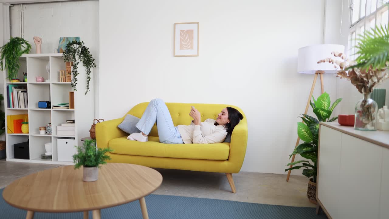 Ung kvinde slapper af på sofaen og bruger smartphone derhjemme