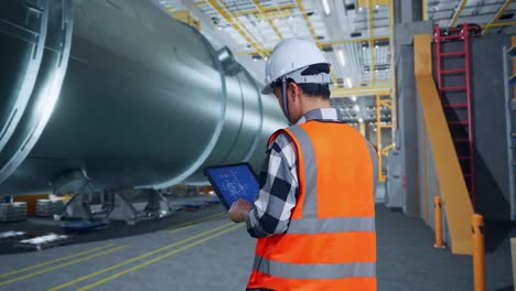 vista trasera de un ingeniero masculino asiático con casco de seguridad trabajando en una tableta con el plan de la turbina eólica en una fábrica de fabricación de tubos