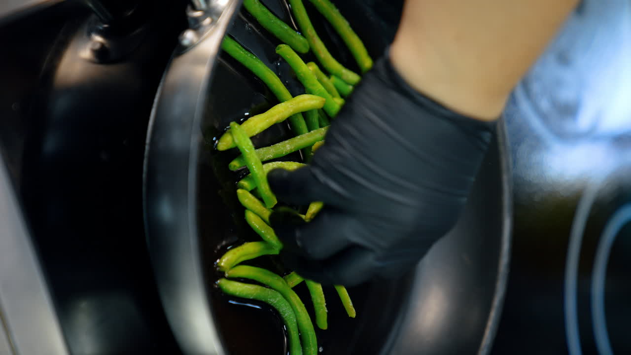 Unrecognized chef's hand in black glove puts the green beans on the frying pan. Cooking veggies close up. Vertical view.