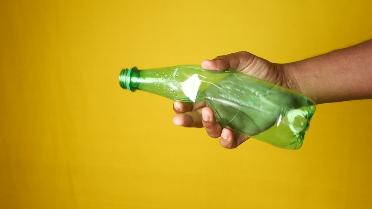 A hand crushing a green plastic bottle against a yellow background