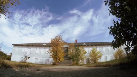 Zeitraffer---Wolken,-Die-An-Einem-Sonnigen-Tag-Hinter-Einem-Alten-Verlassenen-Krankenhaus-In-Einer-Kleinen-Stadt-Mit-Bäumen-Und-Büschen-Um-Den-Hof-Und-Eingänge-Fließen