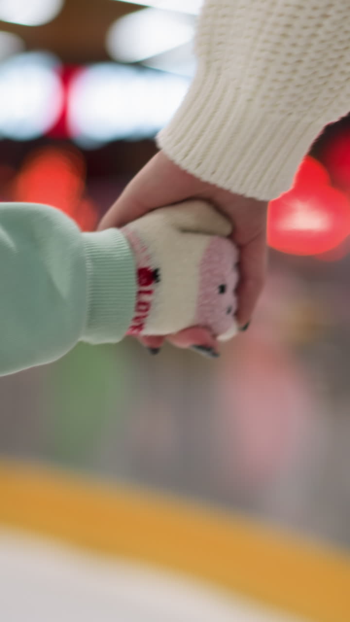 Close-up view of a young girl holding her mom's hand while they skate together on an ice rink, the girl is wearing a mint green outfit, and the mom is dressed in a white sweater