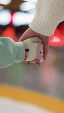 Close-up view of a young girl holding her mom&#039;s hand while they skate together on an ice rink, the girl is wearing a mint green outfit, and the mom is dressed in a white sweater