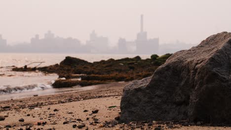 Blick-Auf-Manhattan,-Bedeckt-Mit-Rauch-Von-Waldbränden,-Vom-Strand-Am-East-River-Aus-Gesehen,-Mit-Wellen,-Die-Am-Sandstrand-Krachen,-Und-Moosigen-Felsen-Im-Vordergrund