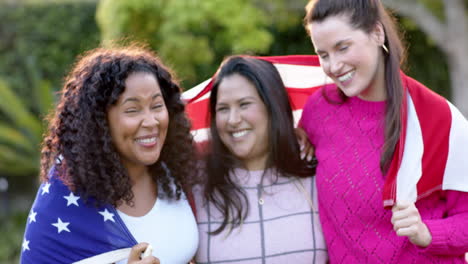 tres felices amigas diversas de pie con la bandera en la espalda en el jardín soleado