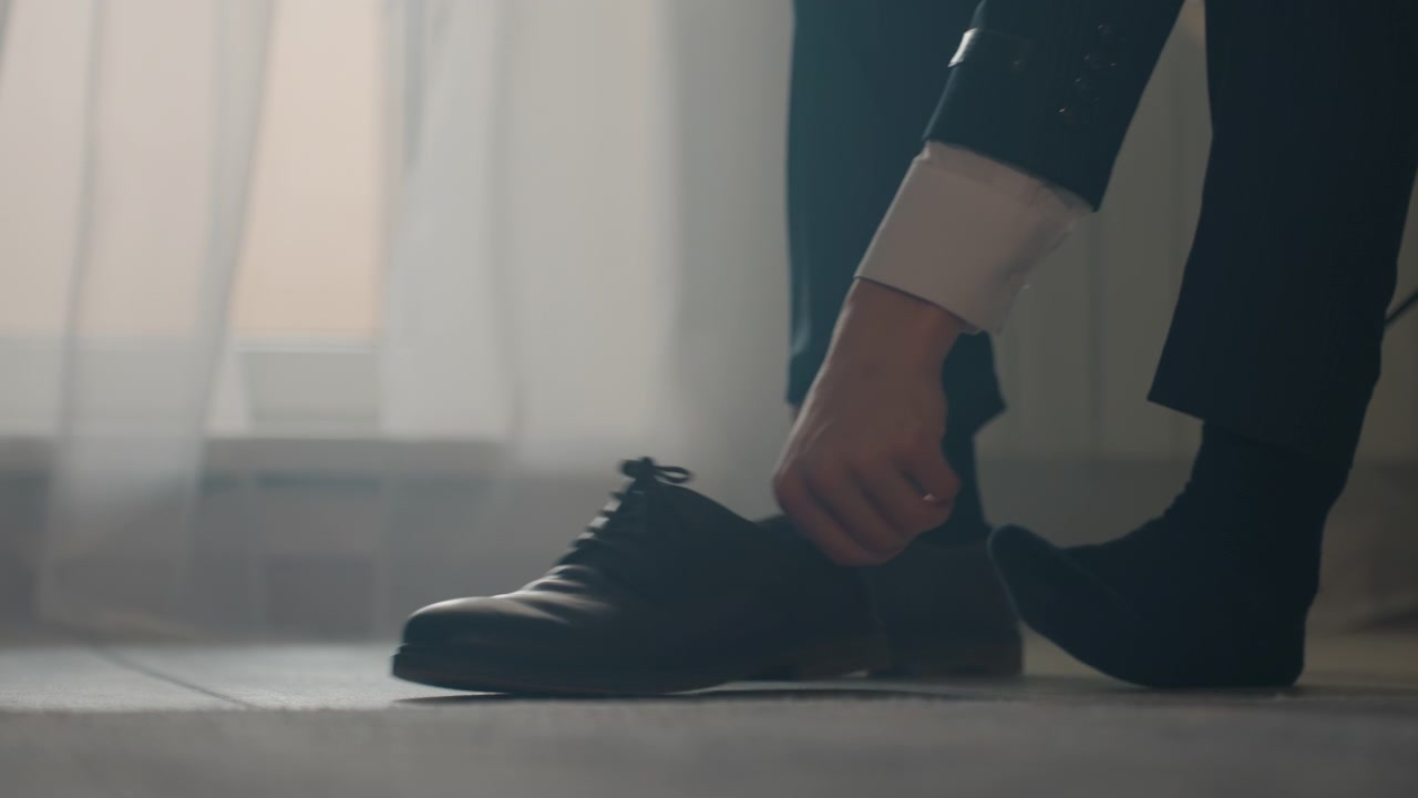 Man tying dress shoes morning light businessman preparing for meeting, polished leather brogues, dark suit trousers, black socks, careful hands adjusting lace, soft diffused window backlight, calm