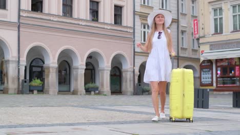 a slender french tourist in a beautiful white dress walks with a yellow suitcase in the center of the old town. tourist woman against the background of beautiful european architecture