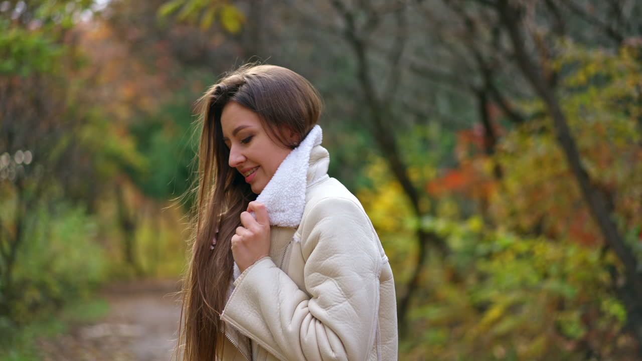 Young woman is hiding in the collar of her white jacket. Long-haired brunette lady walking in the beautiful multi-colored autumn park.