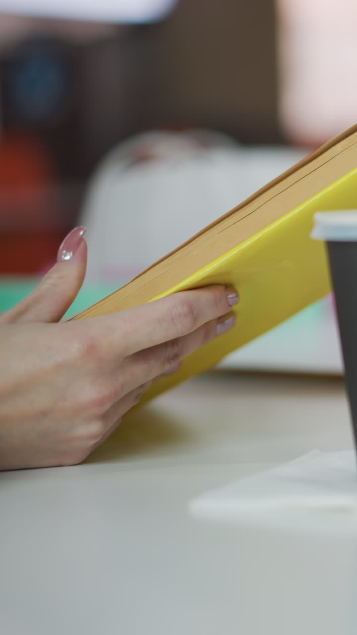primer plano de manos sosteniendo un libro amarillo con uñas pulidas, taza de café en la mesa, y fondo borroso con iluminación cálida y tonos suaves