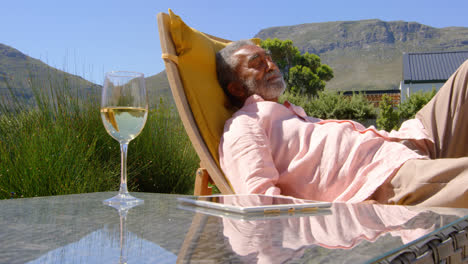 Side-view-of-senior-black-man-relaxing-on-pool-deck-chair-in-back-yard-of-home-4k