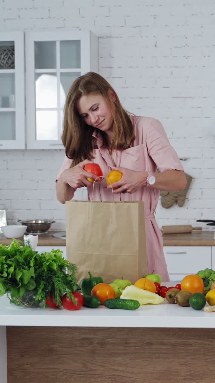 Young happy woman taking out fresh fuitsof a shopping bag. Organic fruit and vegetables in the modern kitchen. Healthy food and dieting concept. Vertical video