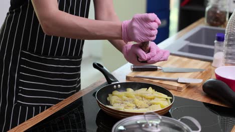 person in gloves and apron seasoning potatoes in a frying pan