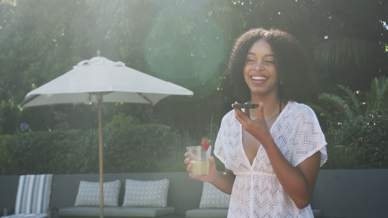 African American woman standing at pool in white dress with phone, answering call, sipping cocktail