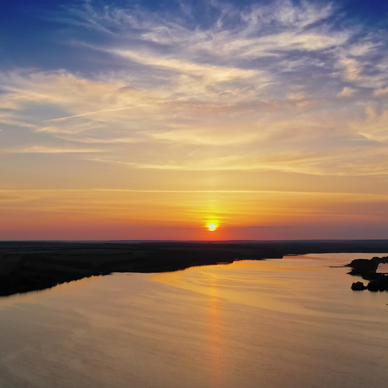 Calm water of the evening lake. Aerial view on the river among natural forests and fields at sunset. Camera moves back.