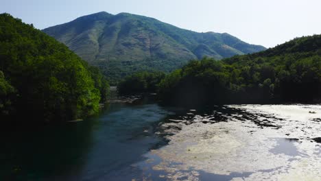 Drone-flying-low-over-blue-water-stream-flowing-amidst-huge-green-mountains