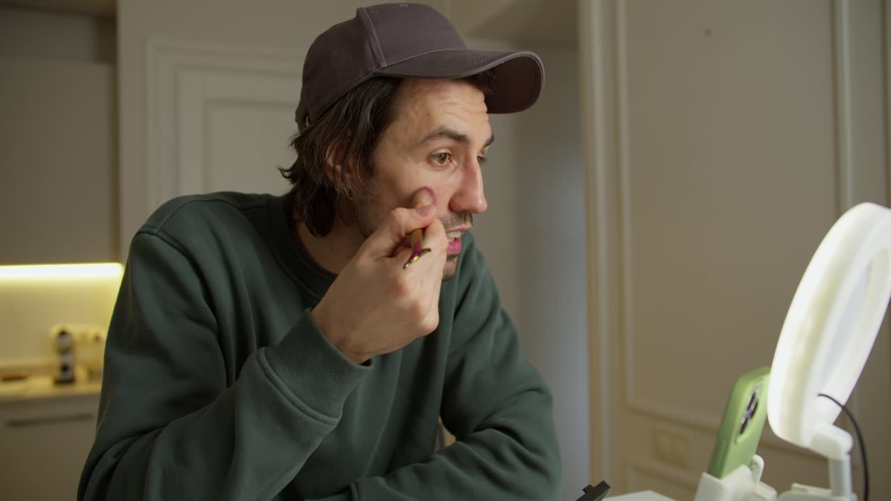 A brunette man in a cap with stubble in a green jacket sits at a table and talks while blogging about how to properly apply powder and other cosmetics to the face in a modern apartment during the day