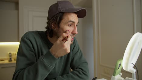 a brunette man in a cap with stubble in a green jacket sits at a table and talks while blogging about how to properly apply powder and other cosmetics to the face in a modern apartment during the day