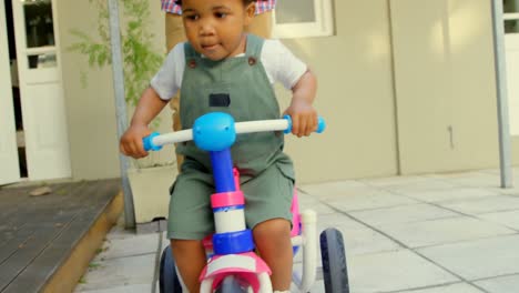 vista frontal de un lindo niño negro montado en un triciclo en el patio trasero de su casa 4k
