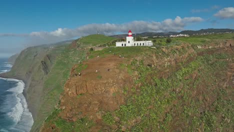 Orbita-Alrededor-De-Los-Turistas-Que-Exploran-Acantilados-Escarpados-Debajo-Del-Faro-Blanco-En-Madeira-Portugal