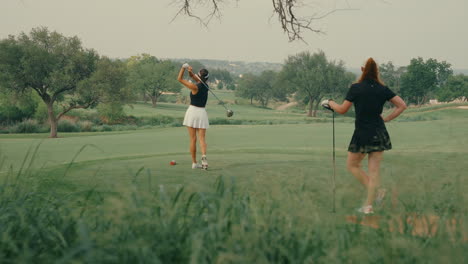 amateur female golfer hits a modest drive off the ladies tees while her playing partner stands behind her, resting on a club. the lush greenery and trees frame the relaxed moment on the course.