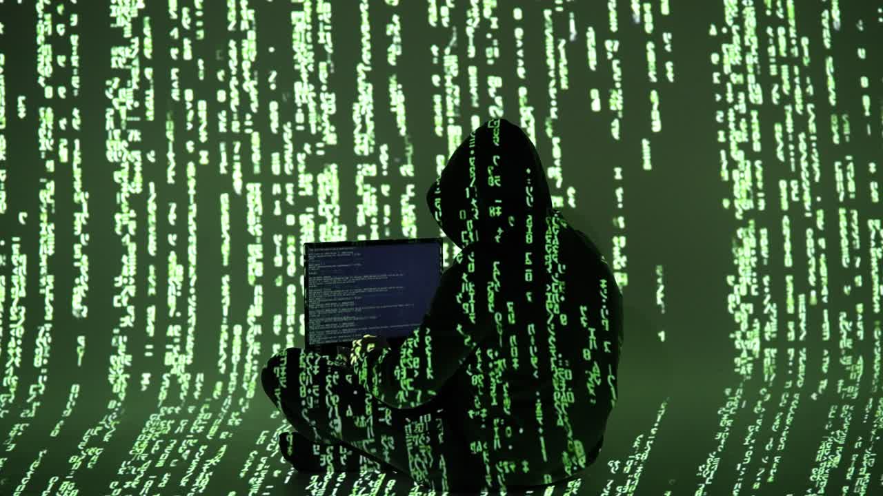 A hooded figure types on a laptop in a dark room filled with green digital rain. The silhouette blends into the matrix-like code, symbolizing stealth, intrusion, and cyber presence
