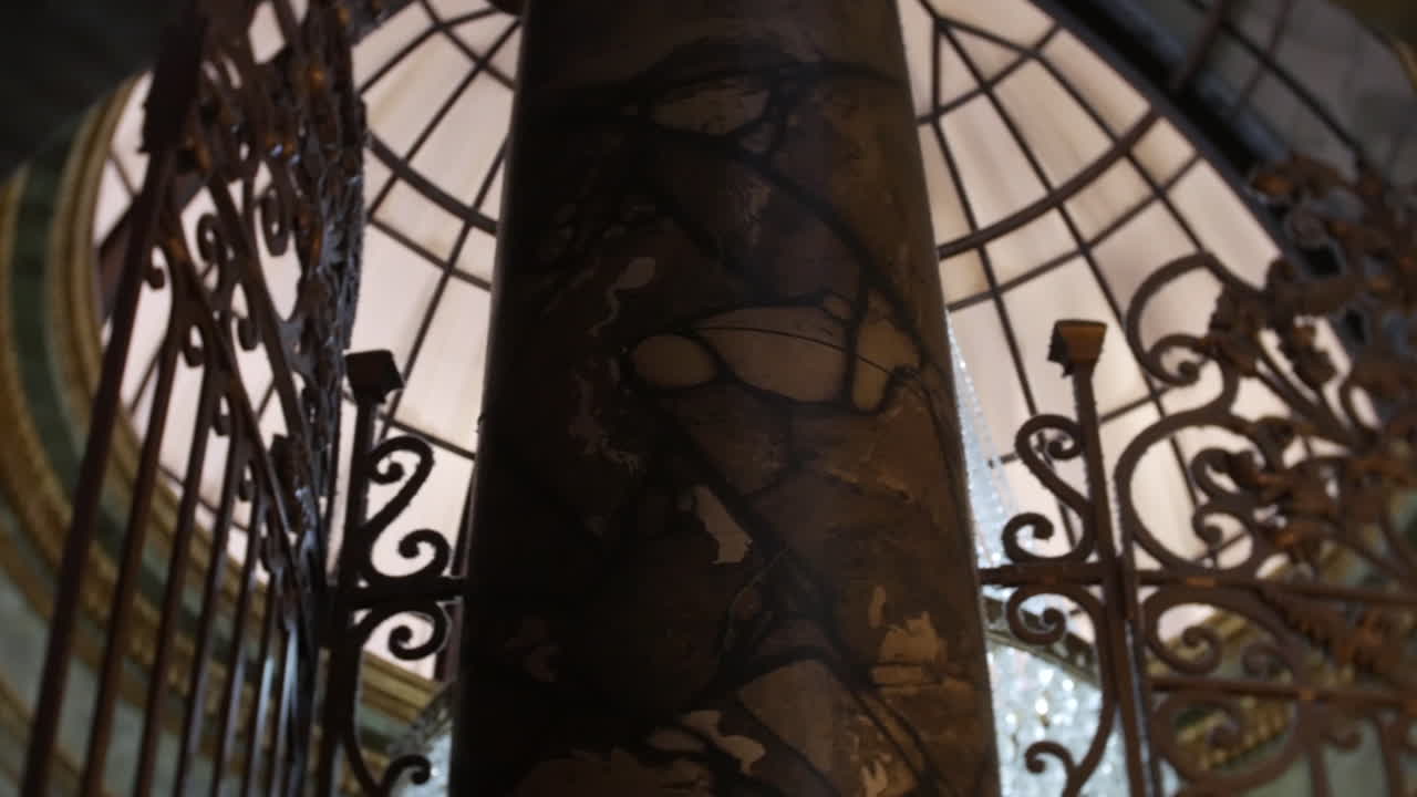 Une balustrade en fer ornée encadre une colonne en marbre texturé sous un plafond en dôme de verre, présentant des détails architecturaux classiques, parfait pour les halls d'hôtels de luxe ou les lieux d'événements haut de gamme.