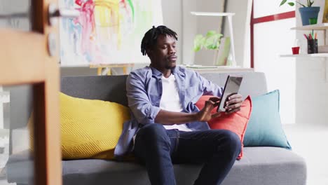 Afroamerikanischer-Männlicher-Künstler,-Der-Ein-Digitales-Tablet-Nutzt,-Während-Er-Auf-Der-Couch-Im-Kunststudio-Sitzt