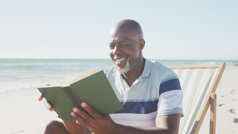 Happy-senior-african-american-man-sitting-on-deck-chair-and-reading-book-at-beach,-in-slow-motion