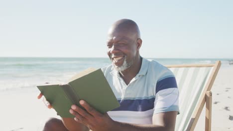 hombre afroamericano mayor feliz sentado en una silla de cubierta y leyendo un libro en la playa, en cámara lenta