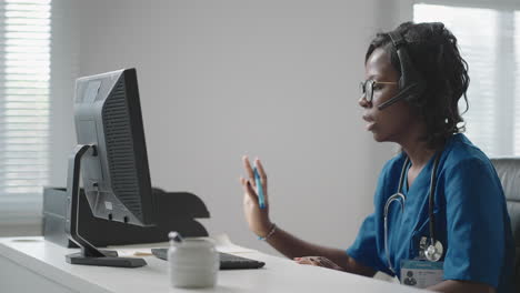 african female medical assistant wears white coat headset video calling distant patient on computer. doctor talking to client using virtual chat computer app. telemedicine remote healthcare