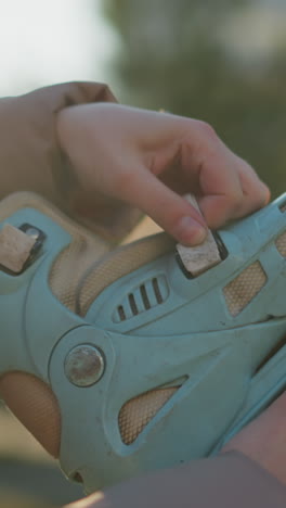 toma de primer plano de una patineta azul claro siendo sostenida y ajustada por una persona en un parque iluminado por el sol. la imagen captura las manos de la persona soltando las correas de la patineta, con un fondo borroso