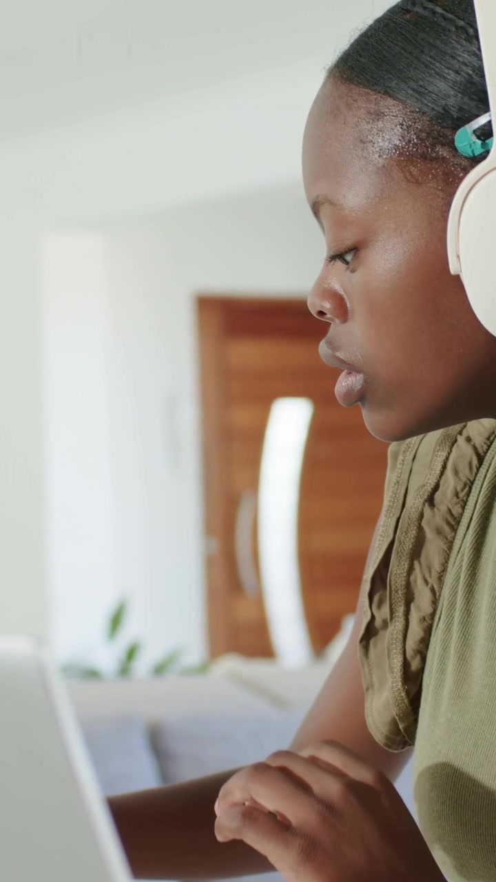 Vertical video: Typing woman in olive top reacting to laptop ping at home, donning headset studying