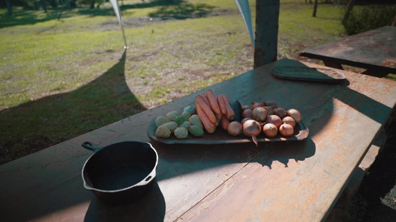 Des carottes, des oignons, de la chayote et une aubergine, des légumes frais sont exposés sur une table en bois rustique à côté d'une poêle en fonte, prêts pour la cuisine en extérieur, plan orbital.