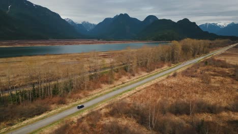 drone-shot-of-a-car-driving-on-a-straight-orad-near-a-Lake-with-mountains-in-the-background,-Pitt-Lake,-Metro-Vancouver,-British-Columbia