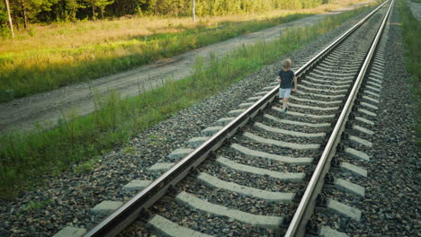 young boy dressed in casual clothes walking alone along rail track in rural setting, passing sign post beside railway with forest and dirt path in background during quiet sunny afternoon