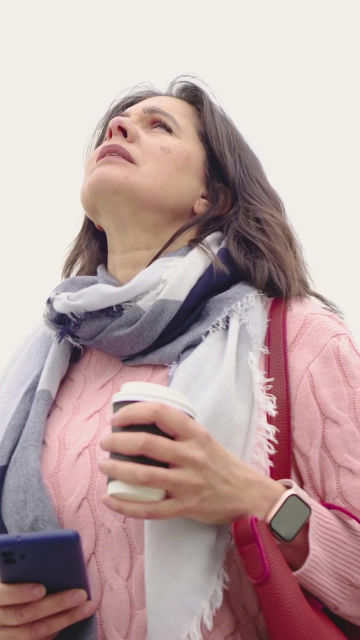 Video verticale: Donna che guarda in alto e scruta il cielo per strada, tenendo un telefono blu e un caffè, con un maglione rosa