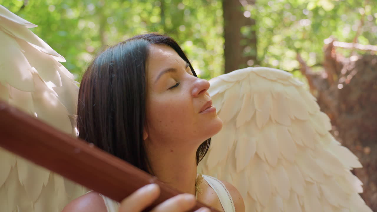 Fairy woman with wide white wings holds wooden staff while gazing upward through sunlight streaming in forest, expression serene and thoughtful, surrounded by glowing leaves